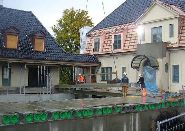 Arbeidere monterer betongelementer med kran under utvidelse av en eldre bygning.