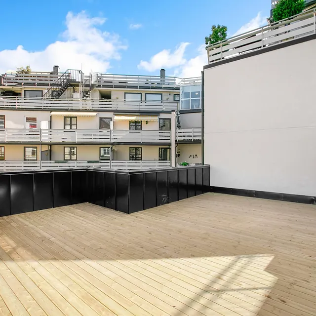 Takterrasse med tregulv, svarte rekkverk og utsikt mot leilighetsbygg.
