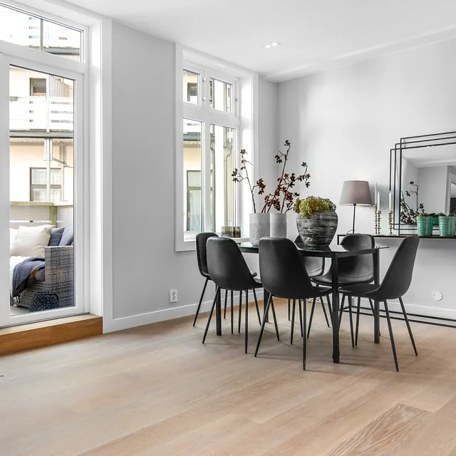 Lys spisestue med svarte stoler og glassbord, store vinduer og dør mot balkong med sittegruppe. Moderne og minimalistisk stil.
