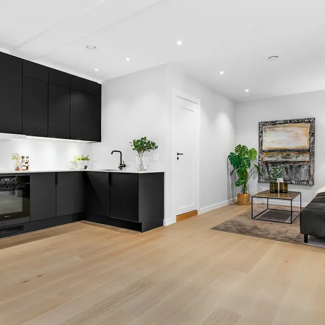Åpen stue og kjøkken med svarte skap, hvite vegger og tregulv. Mørk sofa, kunstverk og grønne planter gir et stilrent uttrykk.